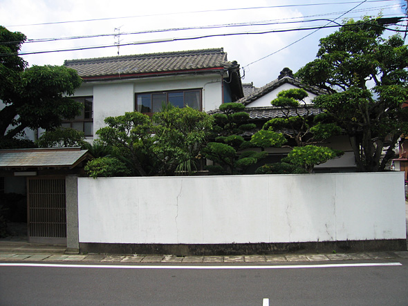 手前に道路があり白い壁と剪定された木々の奥に建つ木造2階建ての建物の写真