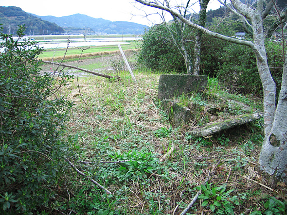 外に田んぼの見える雑木林の入り口に板状の石囲いがある写真