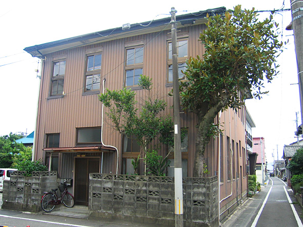 玄関先に自転車が置いてあり、その奥に縦長の窓がついた、2階建ての建物が建っている様子の写真