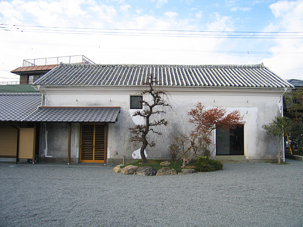 手前に、2本の整備されたような盆栽のようなものが植え付けられており、その奥に平屋の建物が建っている様子の写真