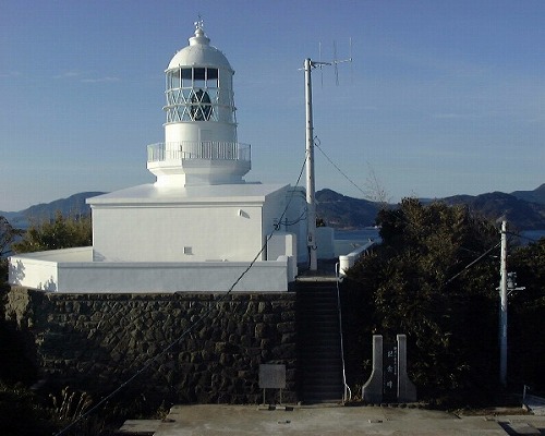 石垣の上に一階建ての建物があり、その上に大きな円柱状の灯台が建っていて、灯台の右横にはアンテナが立ち、その奥に海と丘陵が見えている様子の写真