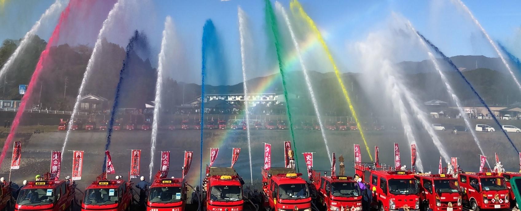 赤色のボディの消防車が横一列に並び、黄色や緑色などの水を一斉放水している 放水は色水を使用して七色のようである。背景には虹もある。