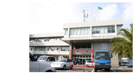 3階建ての日南総合庁舎の外観を駐車場側から写し、ガラス戸の入り口付近を赤い四角で囲っている写真。