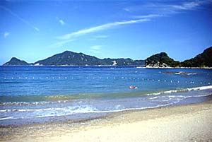大きな岩や山の手前に青い海が広がっており、白い砂浜に波が押し寄せている写真