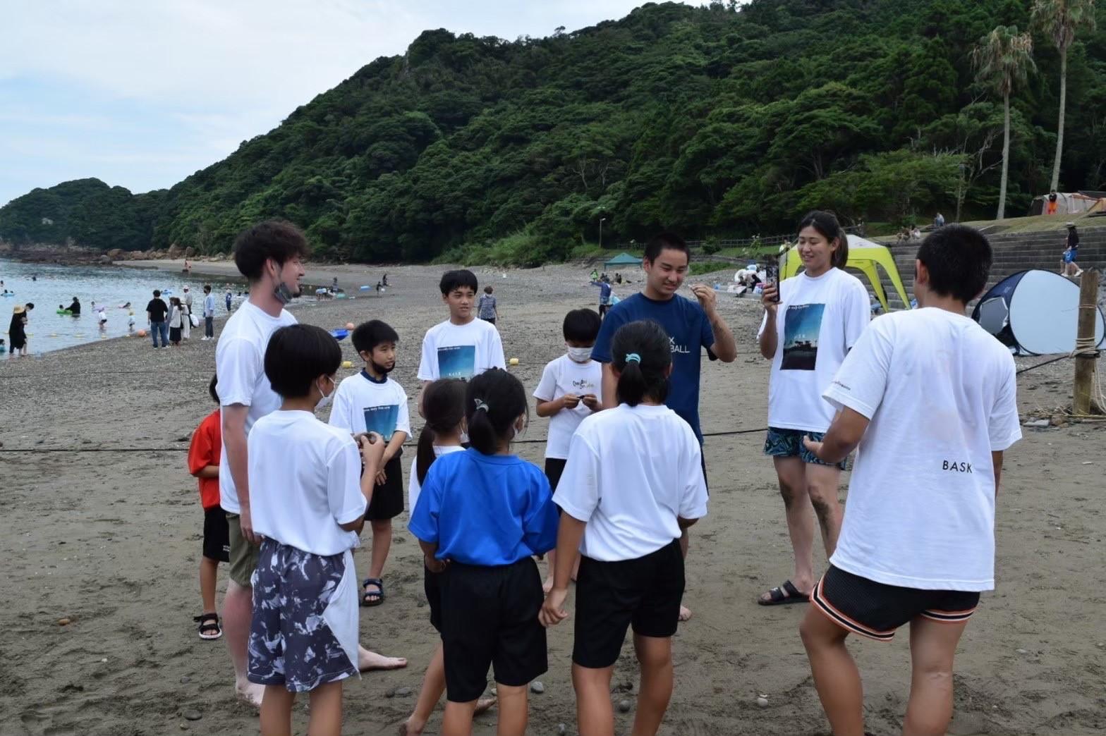 後方に緑の山があるビーチで、井上選手らと子供たちが輪になって話をしている写真