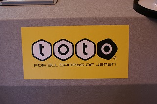 電動式バスケットゴールの土台部分に貼られている、黄色地に描かれた白六角形と黒五角形の中に「toto」の文字が入り、その下に「For all sports of Japan」と記載されている、totoのロゴマークの写真