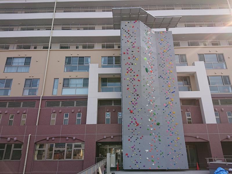 建物の外壁にそって屋外に設置された、建物の5階部分に到達する高さの縦長のグレーの壁に、色とりどりの大小さまざまな石がつけられたリードクライミングウォールの写真
