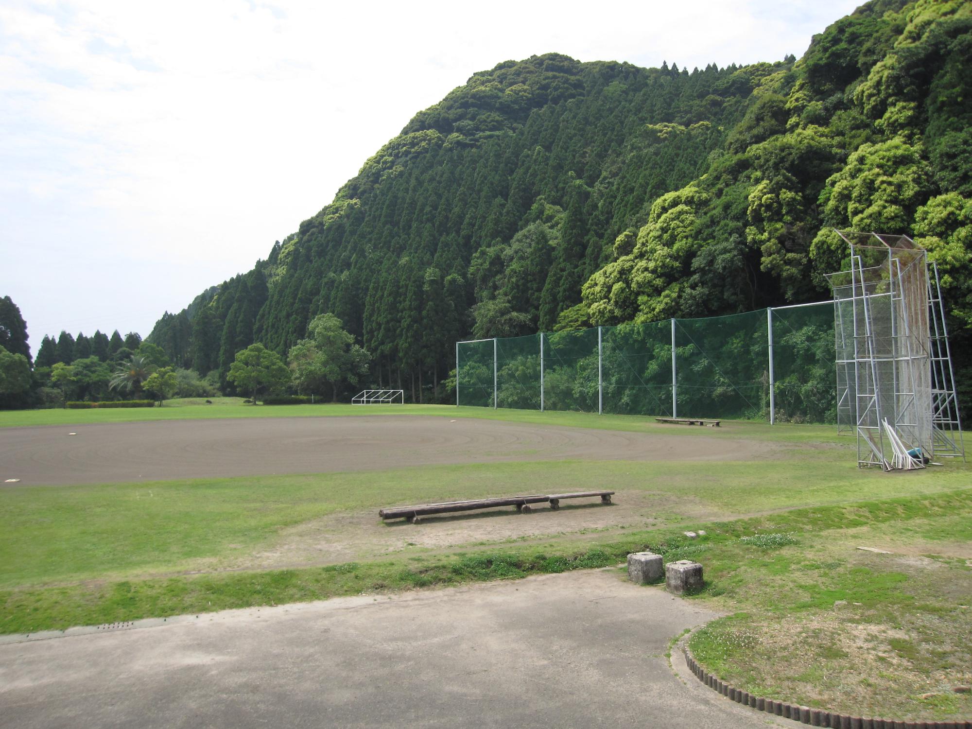 緑の山の手前に広がる、芝生と土の多目的広場にベンチやバックネットがある写真