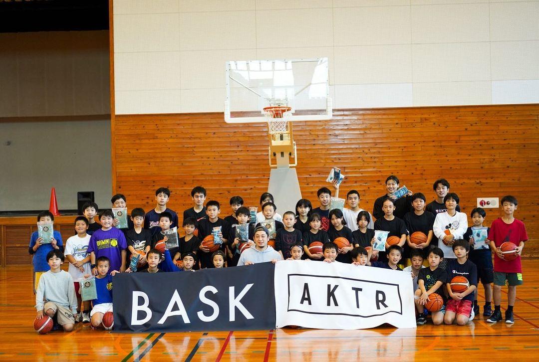 体育館のバスケットゴール前で、黒い生地に白い文字で「BASK」と書かれた横断幕と、白い生地に、黒い四角で囲まれた中に黒で「AKTR」と書かれた横断幕を並べて持ち、丸田氏を中心に、バスケットボールや記念品を手に持った子どもたちが前後3列に並んでいる集合写真