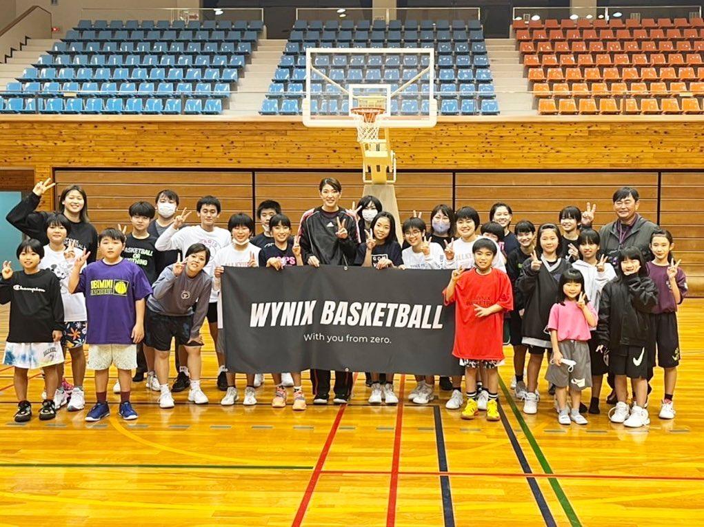 体育館で黒い生地に白い文字で「WYNIX BASKETBALL」と書かれた横断幕を持って、篠原氏を中心に、様々な年齢の子どもたちと関係者が前後3列に並んだ集合写真