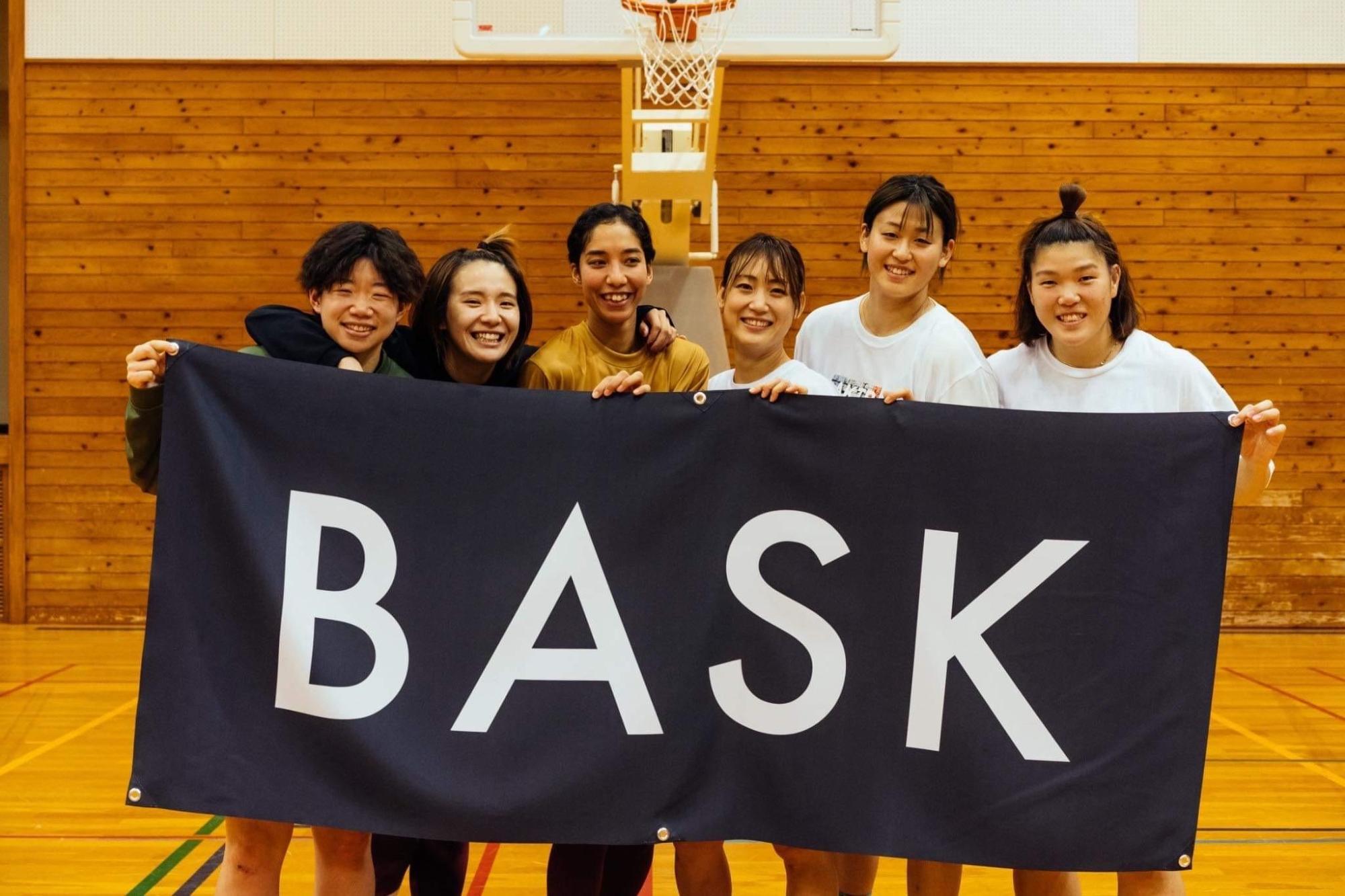 体育館で、黒い生地に白い文字で「BASK」と書かれた横断幕の後ろに、6人の女性が並んで立ち、笑顔で映った記念写真