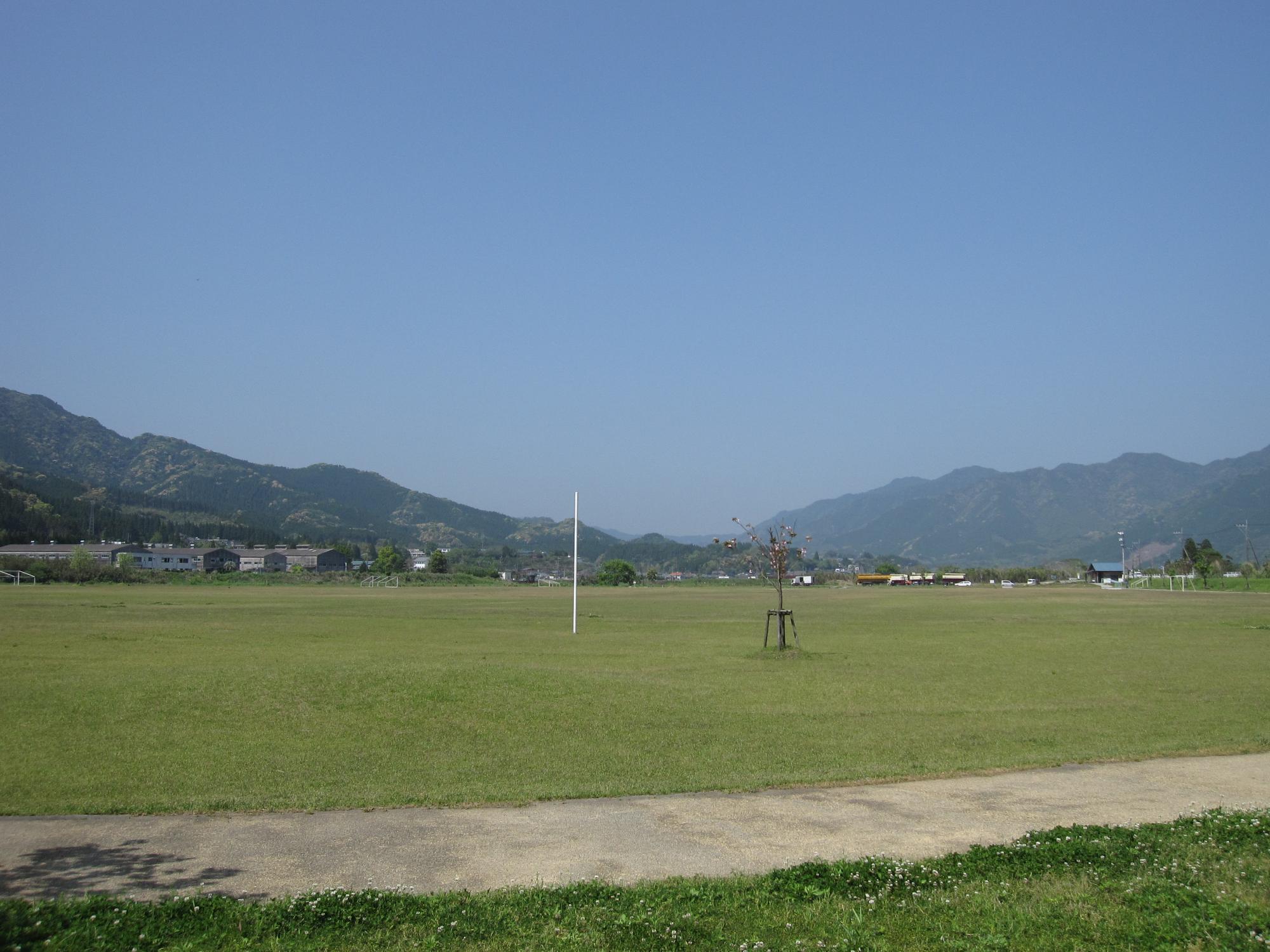 手前に舗装された通路があり、広い芝生の公園の先には山々が見える大藤河川公園ファミリースポーツランドの写真
