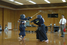 剣道場で、道着や面をつけた二人が竹刀を持って試合をしており、傍らに赤と白の旗を手に持った審判が立っている写真