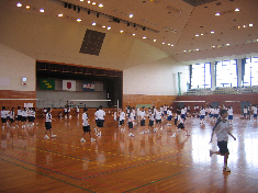 奥の壁中央に国旗などが並んで掲げられた舞台がある天井の高い体育館の中で、体操服を着た沢山の生徒たちが活動している様子の写真