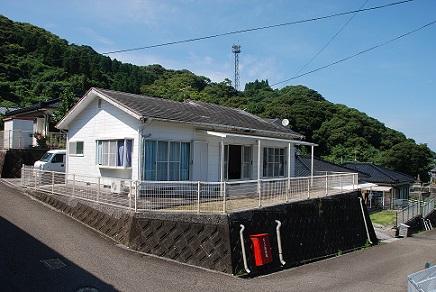 高台に位置しフェンスで囲まれた白壁で平屋建ての建物の移住促進住宅の外観写真