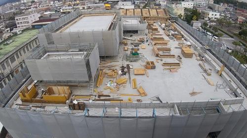 建物全体にグレーのネットがされた足場が組まれ、屋上部分左側に大小2つの四角い建物と沢山の木材が置かれている写真