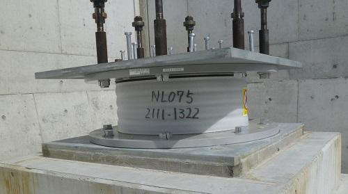 「NL075 2111-1322」と記載された積層ゴム部分が丸い板と正方形の板で挟まれ、上部には黒とシルバーの形状の異なる部品が固定されている天然ゴム系積層ゴム支承である免震装置の写真