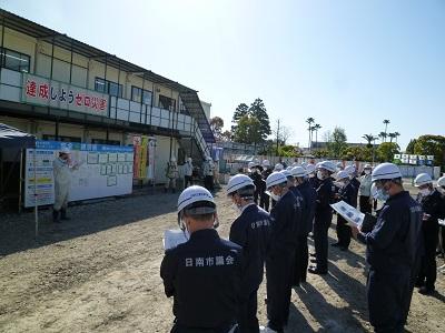 「達成しようゼロ災害」と書かれた横断幕が設置されている二階建ての仮設プレハブの建物の前で話をする男性と、ヘルメットをかぶり「日南市議会」と書かれた作業着を着用した出席者が資料に目を通しながら話を聞いている現場見学会の様子の写真