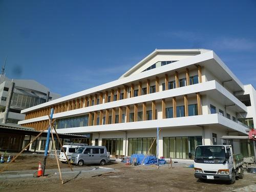 建物前の駐車場部分がまだ舗装されておらず、全体が白を基調とし、正面全面に窓が設置されている3階建て新庁舎の外観写真