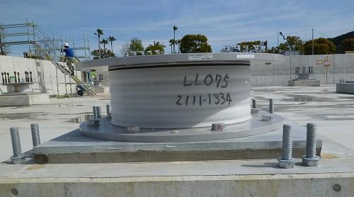 「LL075 2111-1334」と記載された積層ゴムが丸い板と正方形の板で挟まれ、四隅にボルトが設置されプラグ入り積層ゴム支承である免震装置の写真