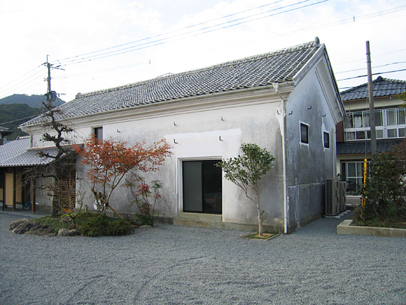 植木が綺麗に手入れされ砂利が敷かれた敷地内に、軒のある入口と、大きな窓が一か所と上部に小さな窓を三か所配置した漆喰塗りと思われる蔵のような建物がある様子の写真