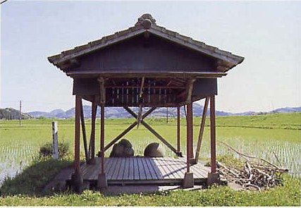 青空の下田圃の脇に祀られている小屋の下にある2つの石の写真