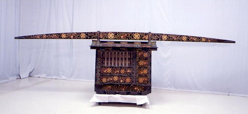 木製で上部に持ち手の棒が取り付けられており、全体に金色で細かな鷹羽紋がデザインされている女乗物の写真