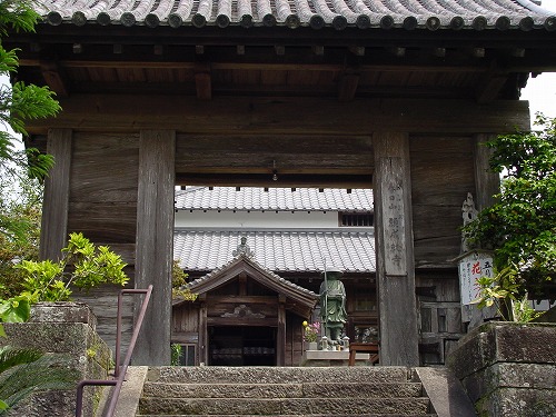 木造で右側に木製の看板が設置された瓦屋根の門が建っており、門の先には願成就寺の建物が見える写真