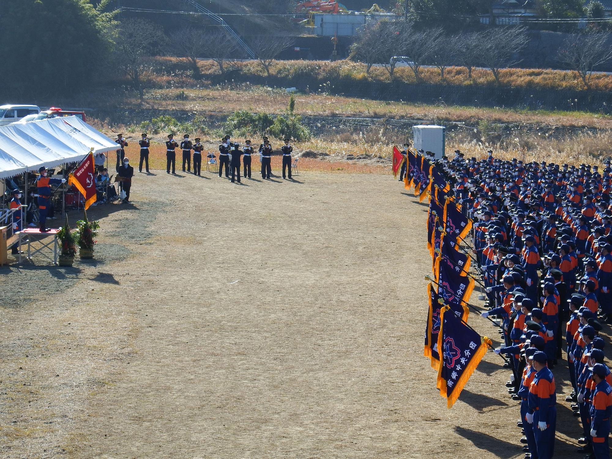 飫肥本町河川敷にて日南市消防団各分団が規則正しくに整列して、各先頭の団員が団旗を掲げている風景