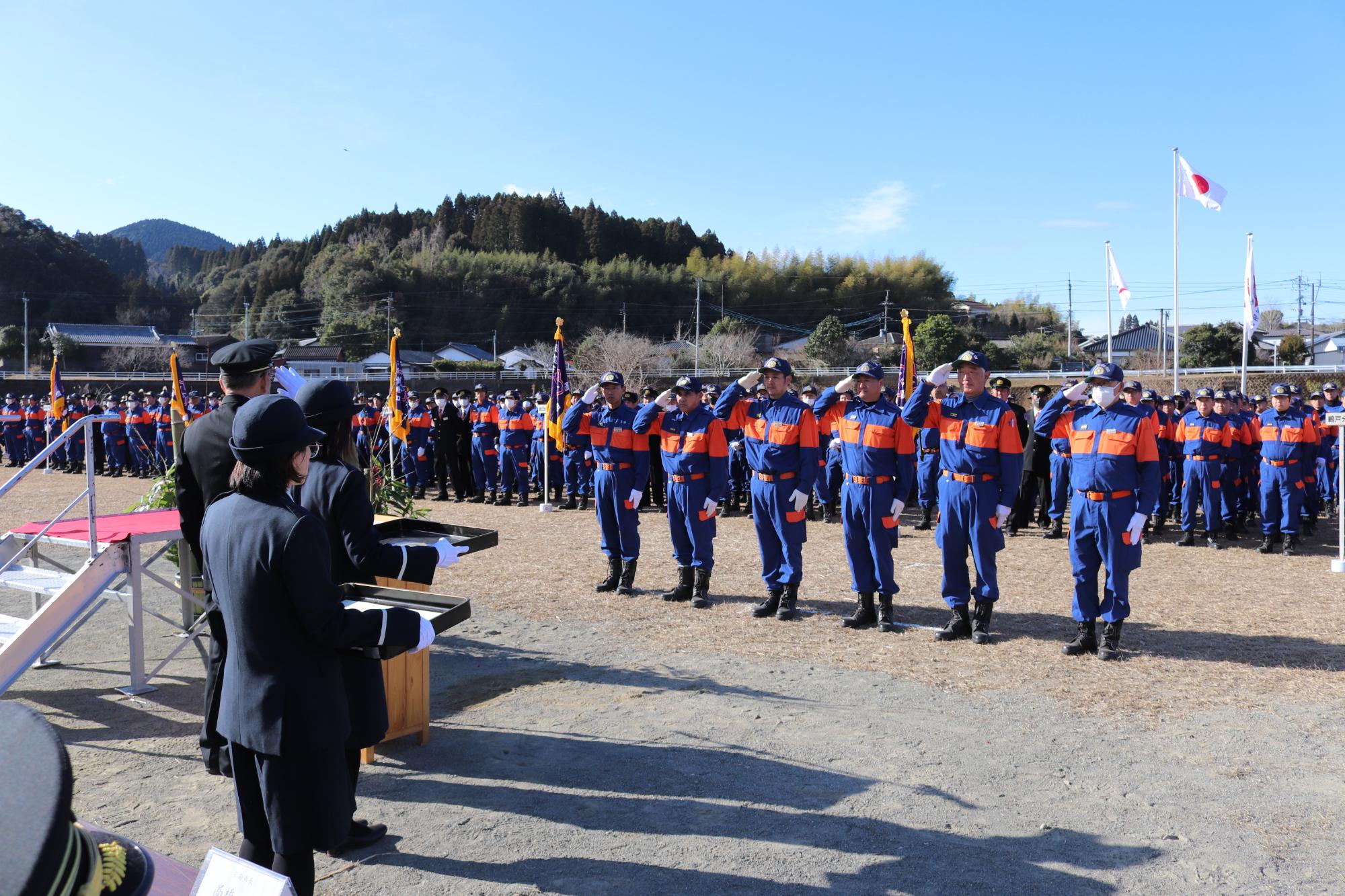 飫肥本町河川敷にて日南市消防団各分団が規則正しくに整列し、表彰者が消防団長に対して敬礼をしている風景
