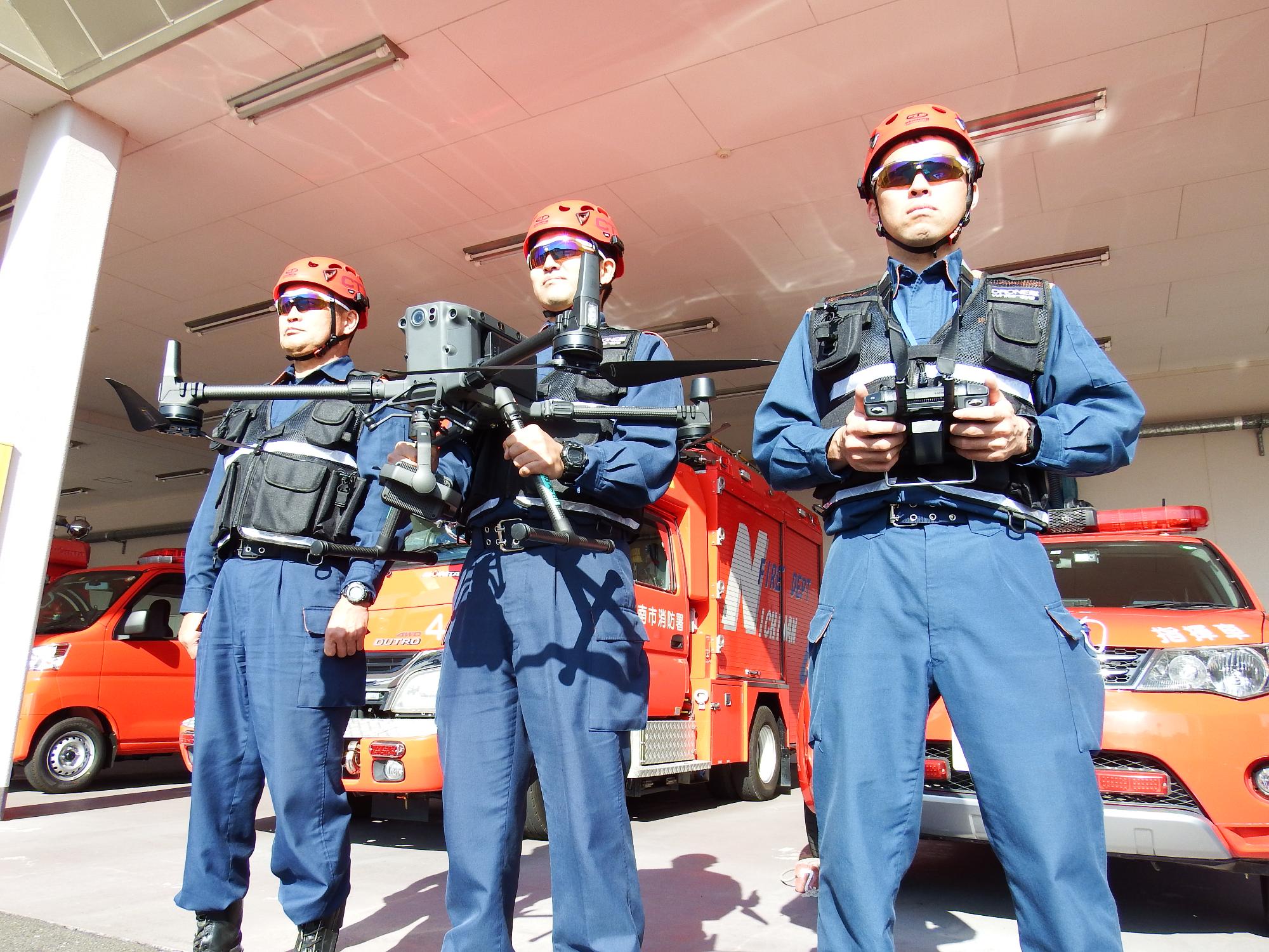 3人の消防職員が、消防車両を背景にドローンの操作訓練を実施している背景です。