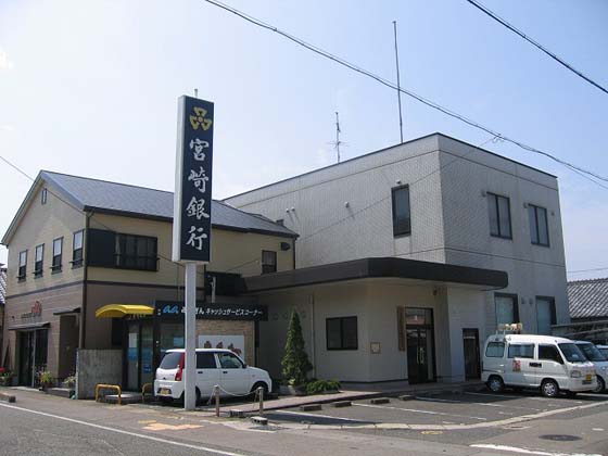 道路沿いに宮崎銀行の青い野立て看板設置されたATMの隣に、隣接された駐車場と2階建ての日南市立大堂津地域交流センター外観写真