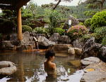 大きな石や植栽のある露天風呂に女性が入浴している後ろ姿の写真
