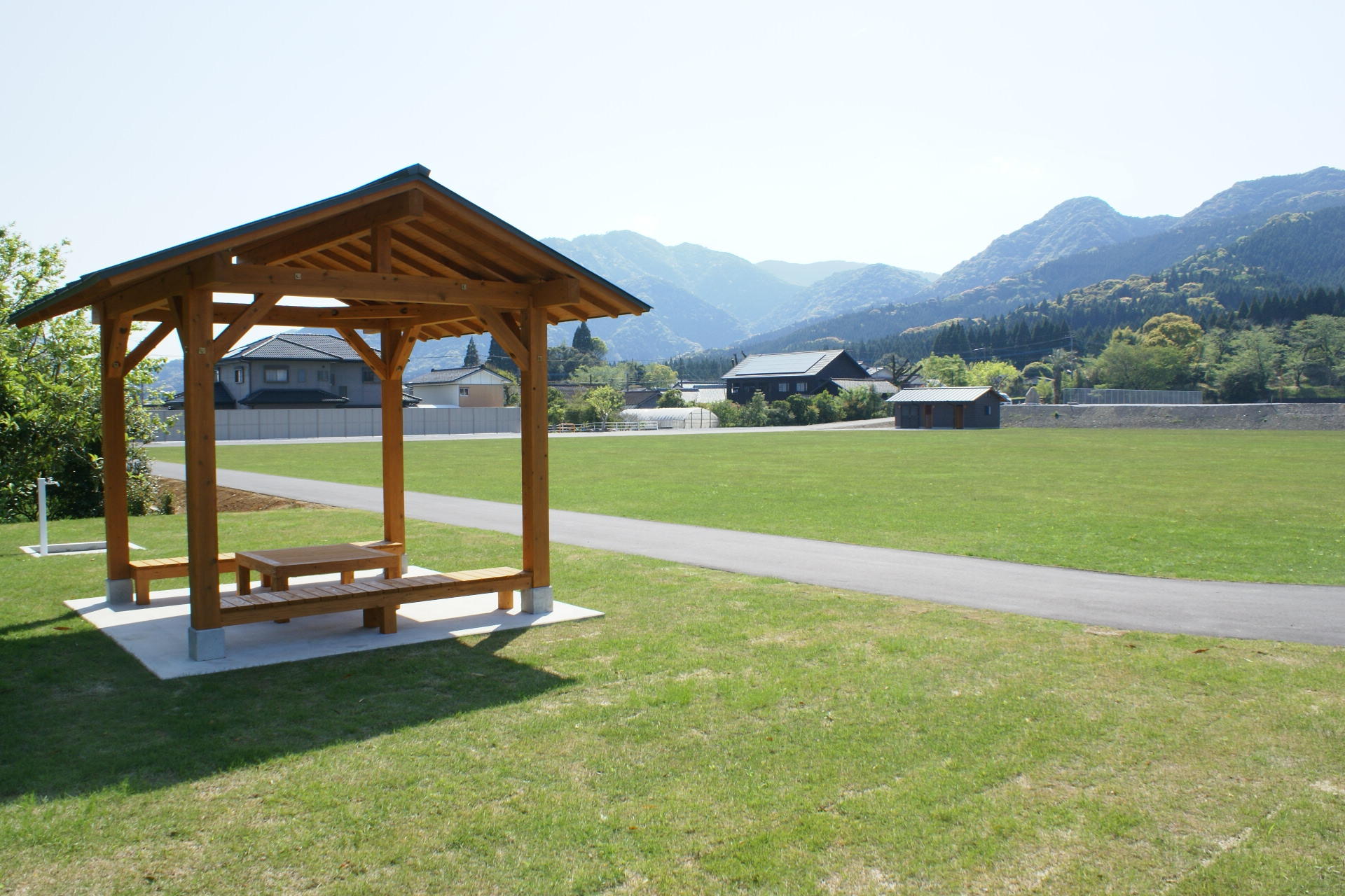 整備された芝生にコンクリートで道が作られており、その手前に木製の東屋が建てられた広々とした多目的芝生広場の写真