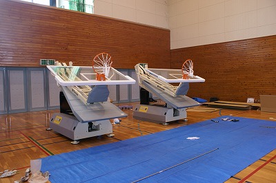 体育館の床に電動式のバスケットゴールが2台置いてあり、その前に青のビニールシートが敷かれている写真