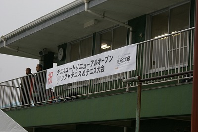 建物の手すりに「祝 テニスコートリニューアルオープン ソフトテニス＆テニス大会」と書かれ、右横にtotoのロゴマークが載った横断幕がかけられており、その左横に男性が2名立っている写真