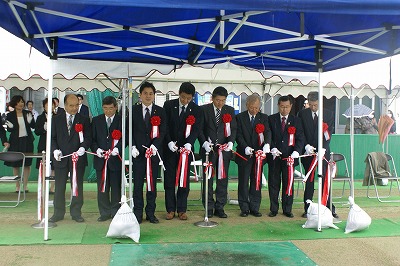 青いテントの下で、胸に赤い花の胸章をつけた8名の男性が横一列に並び、紅白のテープにハサミを入れているテープカットの写真
