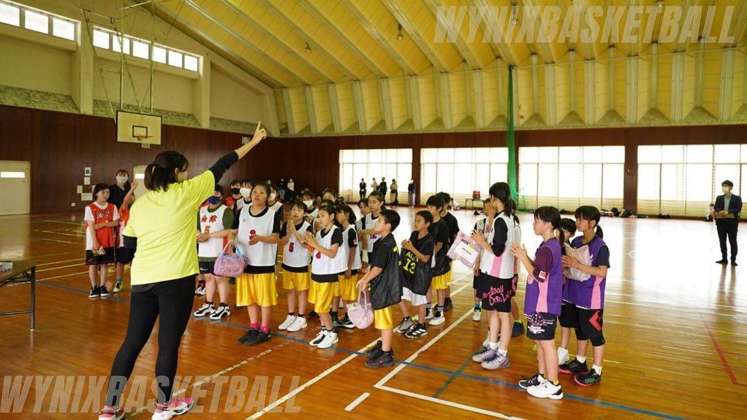 右手人差し指を上に突き上げジェスチャーを交えながら話をする女性と、グループ別に並び話を聞くユニフォームを着用した子どもたちの写真