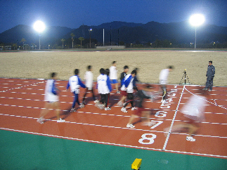 夜間にナイターの電灯で明るく照らされたトラックを走るユニフォームを着た男性たちの写真