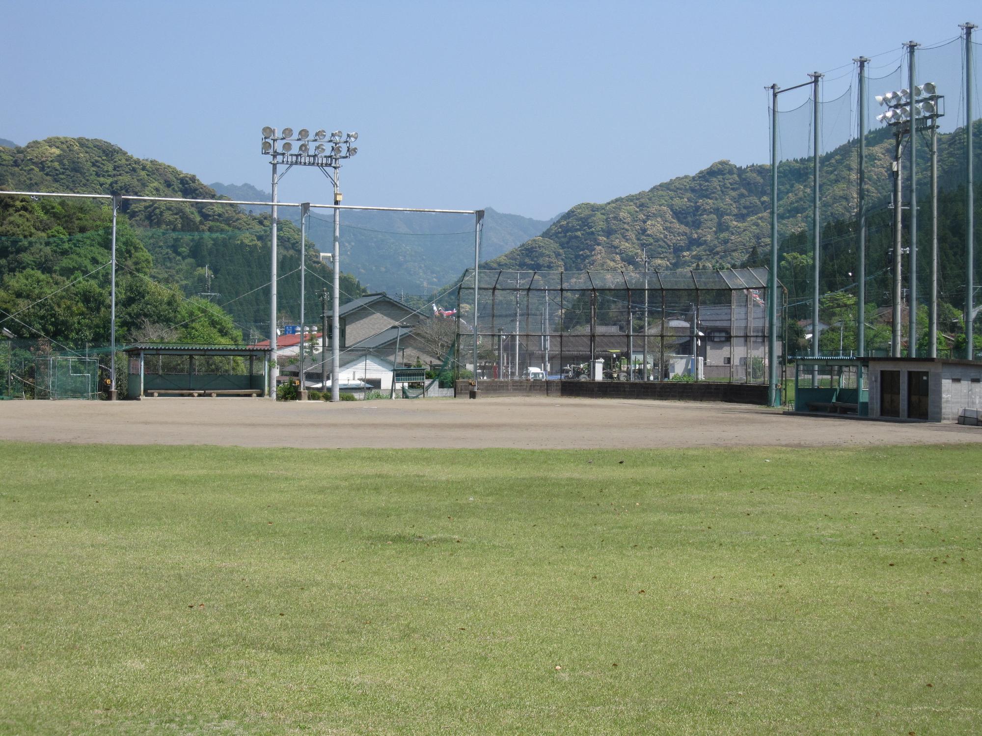 青空の下、周辺の山や家を背景にナイター設備のある北郷坂元球場を天然芝の外野からホームベースの方向にを撮影した写真