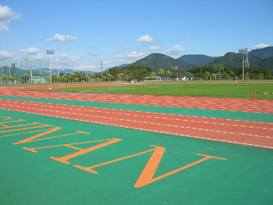 茶色のトラックの内側に芝生のフィールドがあり、外周の緑色床にはオレンジ色でローマ字が書かれている陸上競技場の写真