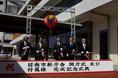 足元に設置されている「祝 日南市新庁舎 開庁式 及び 付属棟 完成記念式典」と書かれた看板の後ろに市長と来賓の方4名が立ち、テープカットが行われた写真