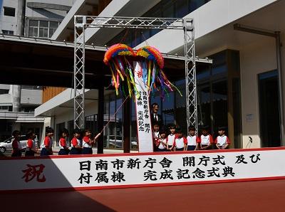 金属の枠組みの上部に下がっているくす玉を、約6名ずつ左右に分かれて並んでいる園児たちがくす玉から伸びたひもを引っ張りくす玉を割っている写真